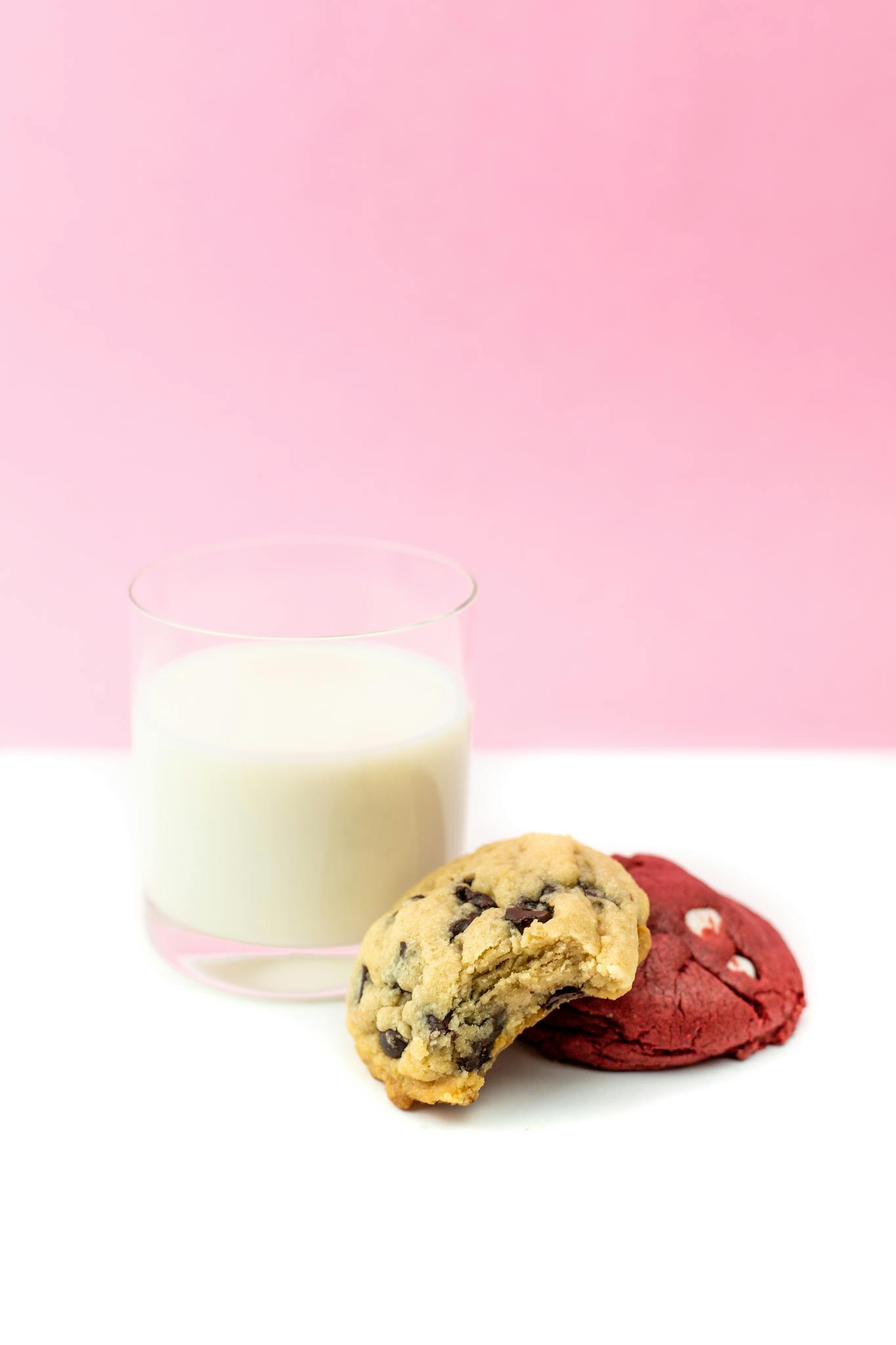 Glass of milk with chocolate chip and red velvet cookies against a pink background.